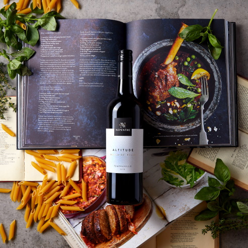 Bottle of nepenthe Altitude Tempranillo across a cooking book, surrounded by basil and pasta